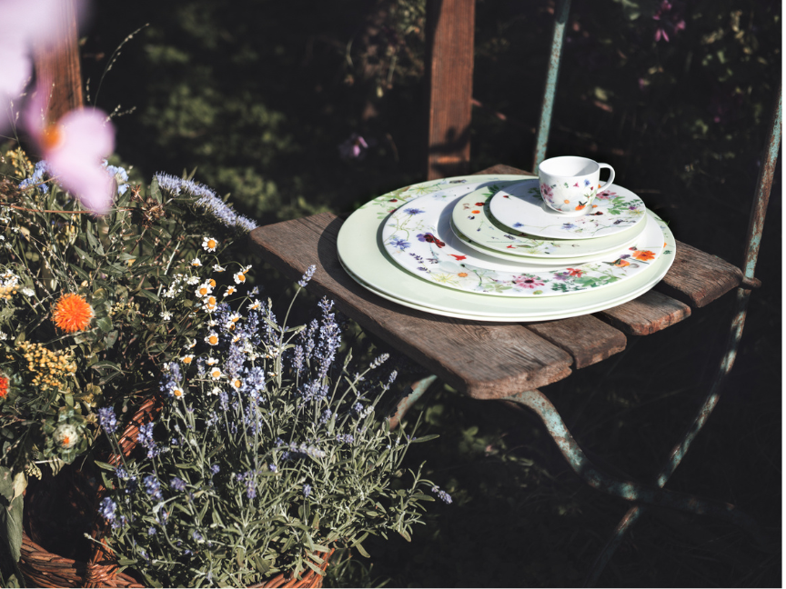 Romantisch gedeckter Gartentisch mit floralen Bone China Porzellan von Rosenthal: Feine Teller und Tasse mit floralem Dekor inmitten bl&uuml;hender Sommerpflanzen.
